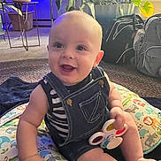 Tatum is registered to the contest to win money with this photo: baby, child, indoor, smiling, denim_overalls, toy, pillow, animal_pattern, plant, carpet, bag, furniture, happy, sitting, person, cute, infant, playful, home, colorful