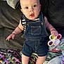 Tatum is registered to the contest to win money with this photo: baby, child, denim_overalls, fidget_spinner, blanket, couch, indoor, curious, sitting, toy, patterned_blanket, infant, clothing, person, face, hand, foot, expression, playful, home