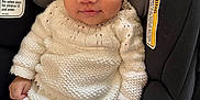 Mariano is registered to the contest to win money with this photo: baby, child, infant, car_seat, knitted_clothing, pacifier, white_outfit, face, eyes, hand, safety_warning, seat_belt, portrait, indoors, cute, smile, young_child, seated, closeup, person
