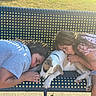 children, dog, puppy, sleeping, cuddling, outdoor, lounge_chair, blanket, pink_dress, blue_shirt, pet, family, companionship, relaxed, sunlight, grass, portrait, animal, comfort, cute