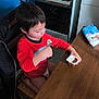child, boy, yogurt_cup, spoon, kitchen, stove, frying_pan, table, chair, red_shirt, jackets, wooden_table, indoor, snack, eating, kneeling, short_hair, tile_floor, packet, counter