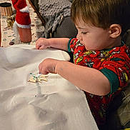 Alex Allred is registered to the contest to win money with this photo: toddler, child, pajamas, christmas, holiday, ornament, wooden, marker, table, blanket, couch, decorations, red_clothing, indoor, crafting, face, person, furniture, holiday_decor, activity