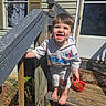 child, toddler, smiling, outdoor, wooden_steps, porch, sunlight, barefoot, clothing, spider_man, red_bowl, pet_food, doormat, ghost_pattern, railing, house, window, door, happy, person