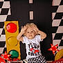 checkered_flag, checkered_pattern, child, curly_hair, decor, green_light, hands_up, indoor, playful, racing_theme, red_light, red_star_light, seat, smiling, toddler, toy_car, traffic_light, white_tshirt, yellow_light, yellow_tire