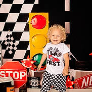Caleb is registered to the contest to win money with this photo: black_shoes, checkered_flag, checkered_shorts, child, curly_hair, decor, happy, indoor, playful, race_theme, racing_outfit, red_star_light, route_66_sign, smiling, standing, stop_sign, toddler, toy_car, traffic_light, white_shirt