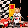 black_shoes, checkered_flag, checkered_shorts, child, curly_hair, decor, happy, indoor, playful, race_theme, racing_outfit, red_star_light, route_66_sign, smiling, standing, stop_sign, toddler, toy_car, traffic_light, white_shirt