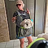 person, dog, pet_carrier, mirror_selfie, sunglasses, cap, casual_clothing, elevator, tile_floor, marble_wall, indoor, reflection, hand_gesture, peace_sign, shorts, white_shoes, fluffy_dog, black_tshirt, phone, cute