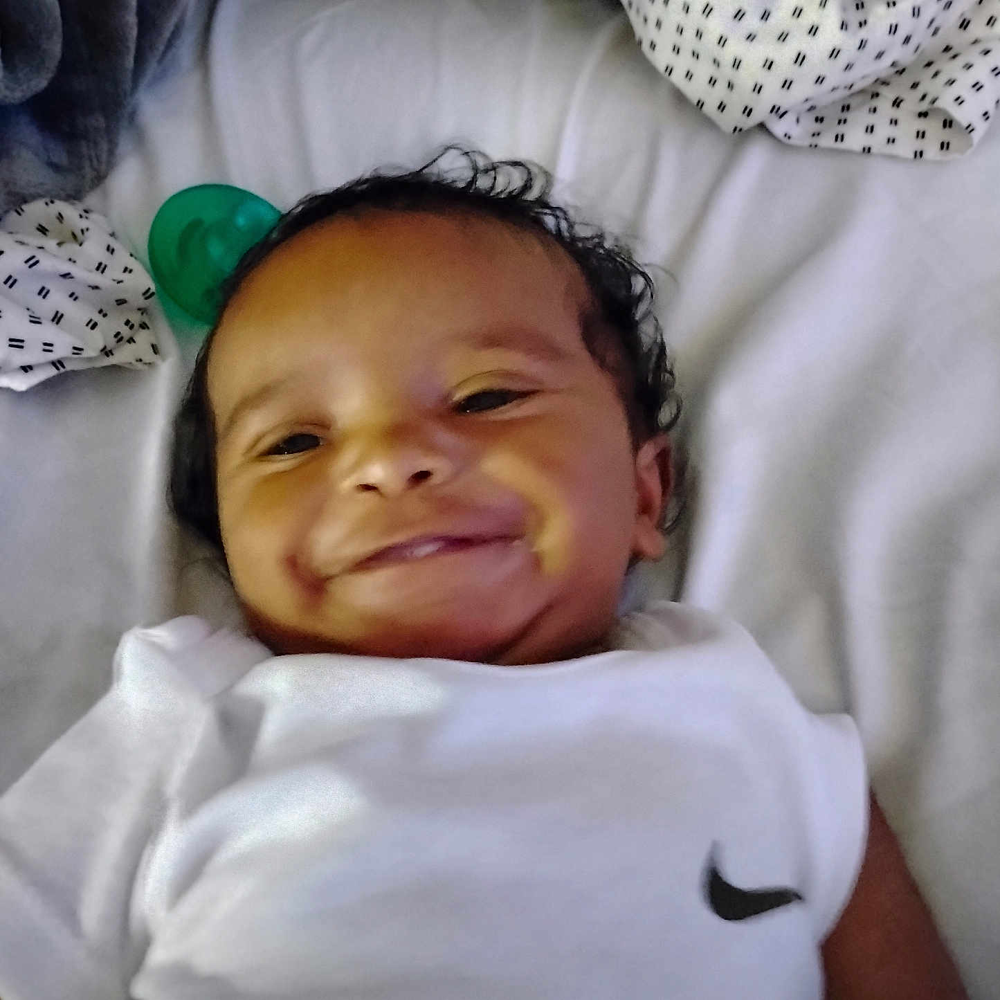 Dedrick joined the competition — help win amazing prizes! baby, bed, blanket, child, cozy, curly_hair, cute, face, happy, indoor, infant, lying_down, nike_logo, pacifier, person, portrait, resting, smiling, soft_texture, white_clothing