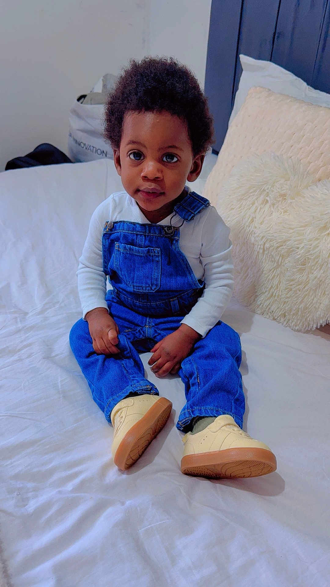 Giovanni a rejoint le concours — aidez-le/la à gagner de superbes lots ! toddler, child, denim_overalls, white_shirt, sneakers, bed, pillow, indoor, portrait, cute, curly_hair, young_child, sitting, cozy, casual_clothing, person, face, eyes, hands, home