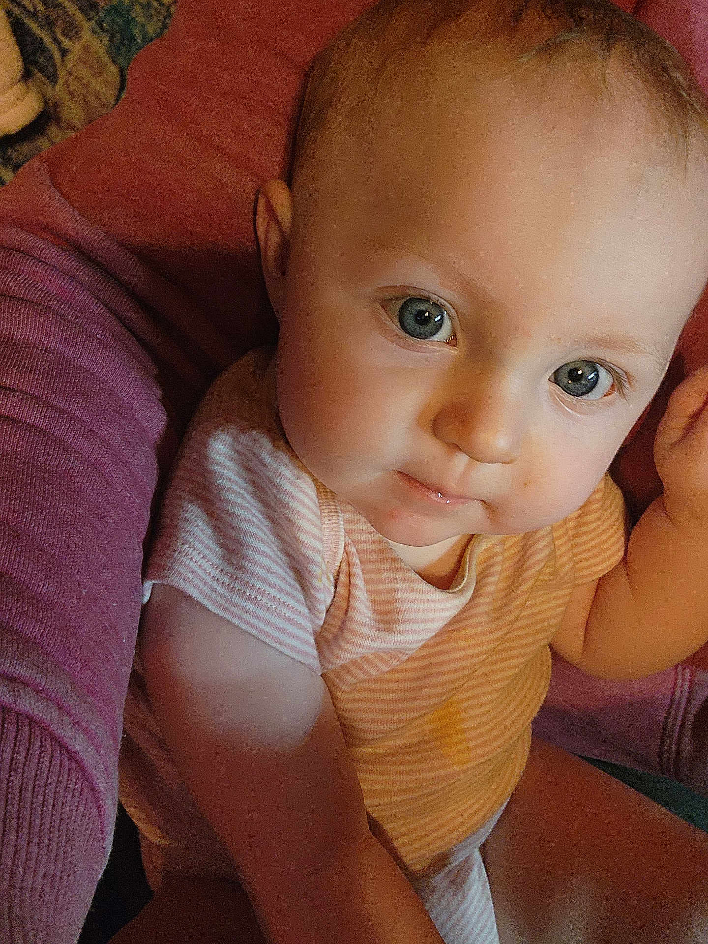 Aubrey joined the competition — help win amazing prizes! baby, infant, face, blue_eyes, onesie, striped_clothing, portrait, close_up, smile, hand, arm, parent_arm, sofa, fabric, soft_texture, skin, eyelashes, indoor, warm_lighting, cozy