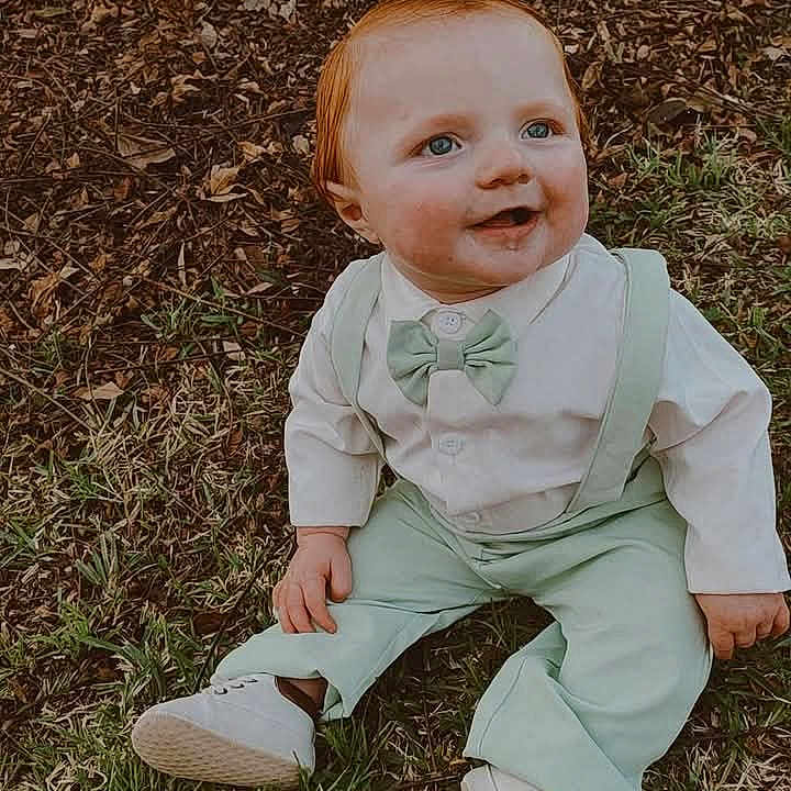 Zayden is registered to the contest to win money with this photo: baby, blue_eyes, bow_tie, casual_clothing, child, cute, daylight, fall_leaves, grass, happy, nature, outdoor, portrait, red_hair, sitting, smiling, suspenders, tree, white_shoes, young_child