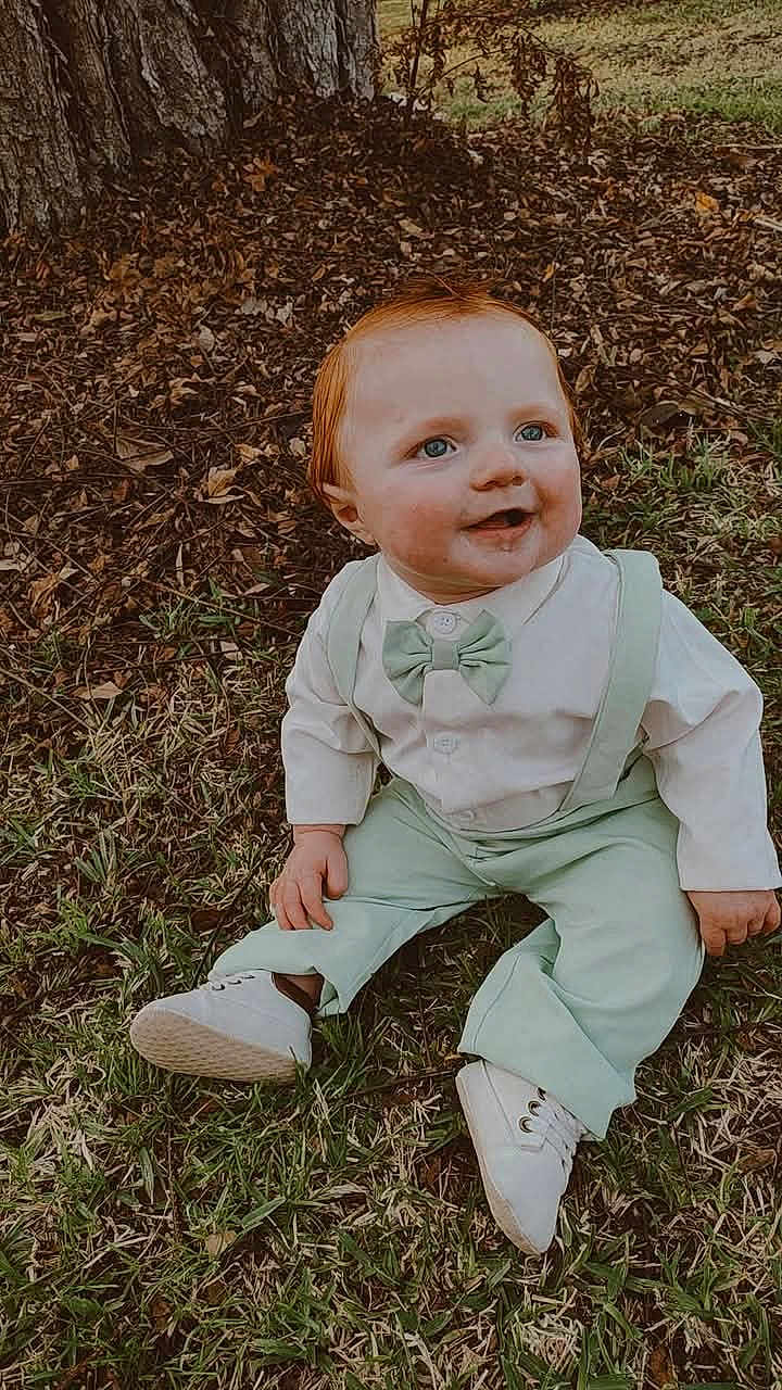 Zayden is registered to the contest to win money with this photo: baby, child, grass, tree, outdoor, smiling, happy, blue_eyes, red_hair, suspenders, bow_tie, white_shoes, cute, nature, fall_leaves, sitting, portrait, young_child, casual_clothing, daylight