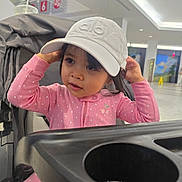Arya is registered to the contest to win money with this photo: toddler, child, pink_clothing, hat, cap, stroller, indoor, curious, face, portrait, polka_dot, drink, cup, modern, public_space, expression, person, young_child, casual, seated