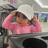 toddler, child, pink_clothing, hat, cap, stroller, indoor, curious, face, portrait, polka_dot, drink, cup, modern, public_space, expression, person, young_child, casual, seated