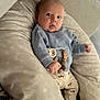 baby, infant, cushion, pillow, clothing, blue_sweater, beige_pants, wooden_beads, patterned_fabric, face, eyes, hand, sock, indoors, soft_lighting, cute, comfort, person, child, portrait