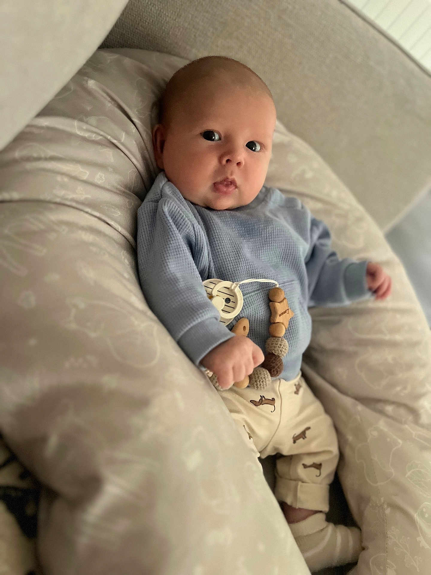Lennon participe au concours pour gagner de l'argent avec cette photo : baby, infant, cushion, pillow, clothing, blue_sweater, beige_pants, wooden_beads, patterned_fabric, face, eyes, hand, sock, indoors, soft_lighting, cute, comfort, person, child, portrait