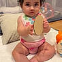 baby, child, infant, curly_hair, red_bow, bib, toy, wooden_toy, sitting, blanket, cushion, indoor, floor, playpen, cute, pink_clothing, expression, face, person, home