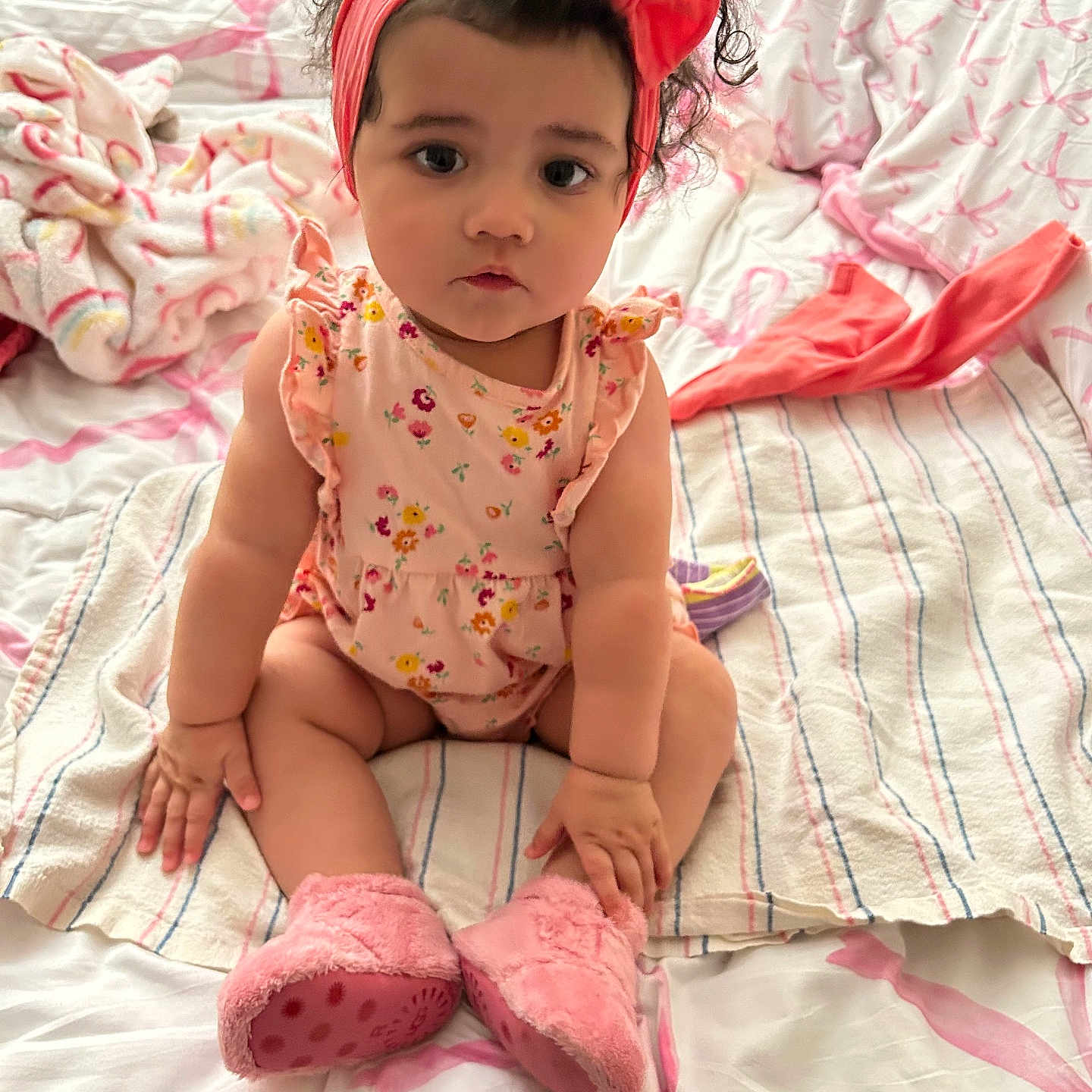Yazmine is registered to the contest to win money with this photo: baby, bed, blanket, boots, child, clothing, cozy, curly_hair, cute, floral_pattern, headband, indoors, person, pink, portrait, sitting, soft, striped_blanket, toddler, young_child