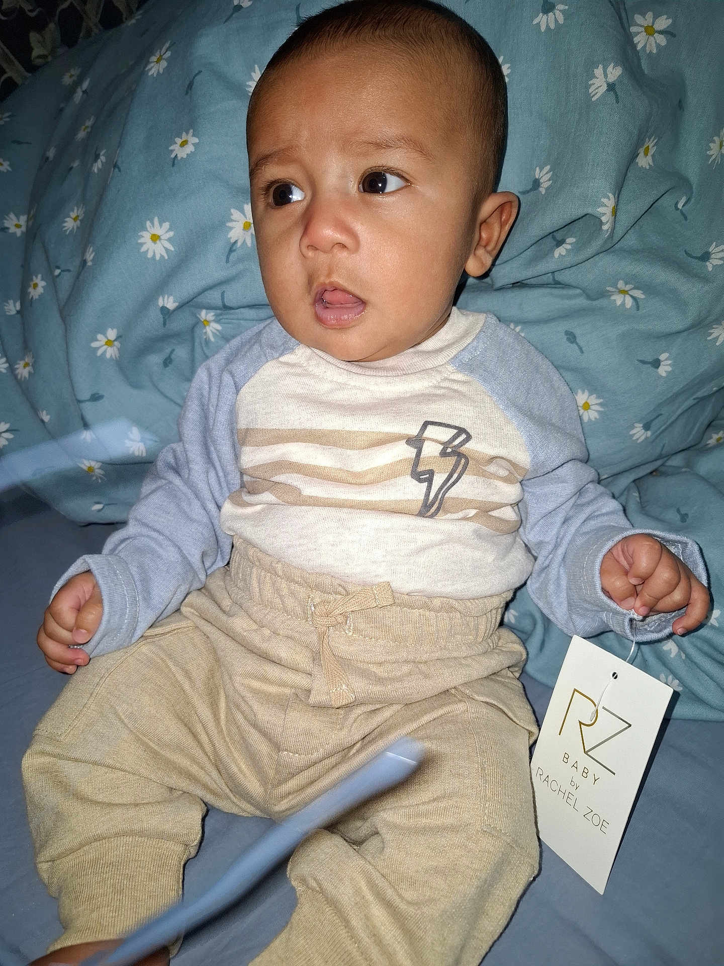 Turhan joined the competition — help win amazing prizes! baby, child, clothing, pants, shirt, tag, bed, blue, floral, sheet, person, face, hand, sitting, curious, expression, indoor, cozy, lightning_bolt, apparel