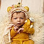 baby, infant, teddy_bear, knitwear, yellow_clothing, bonnet, floral, bear_ears, blanket, fur_texture, wooden_floor, cute, portrait, child, newborn, soft_lighting, indoors, expression, hand_position, cozy