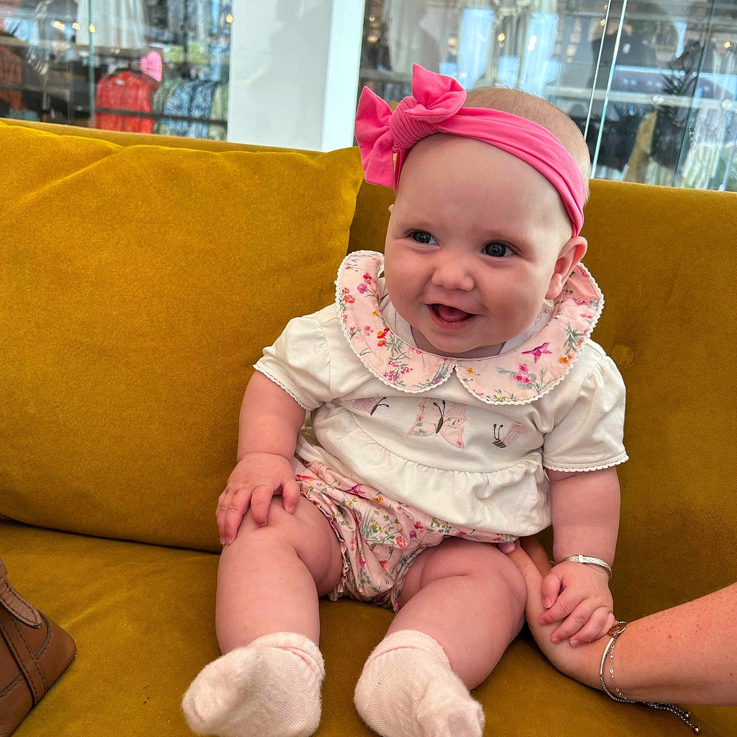 Elaya joined the competition — help win amazing prizes! adult_hand, baby, child, clothing_store, couch, floral_clothing, glass, hand_holding, happy, headband, indoor, infant, pink_headband, reflection, sitting, smiling, socks, store, white_shirt, yellow_couch