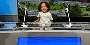 Giovanni participe au concours pour gagner de l'argent avec cette photo : child, smile, curly_hair, microphone, news_desk, television_studio, blue_screen, cityscape, screen, technology, indoor, happy, person, broadcast, media, studio_lighting, desk, young, portrait, background