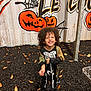 child, smiling, halloween, costume, skeleton, jack_o_lantern, pumpkin, gravel, autumn_leaves, outdoor, curly_hair, black_clothing, playful, decorated_wall, spider_web, footwear, joyful, festive, night, person