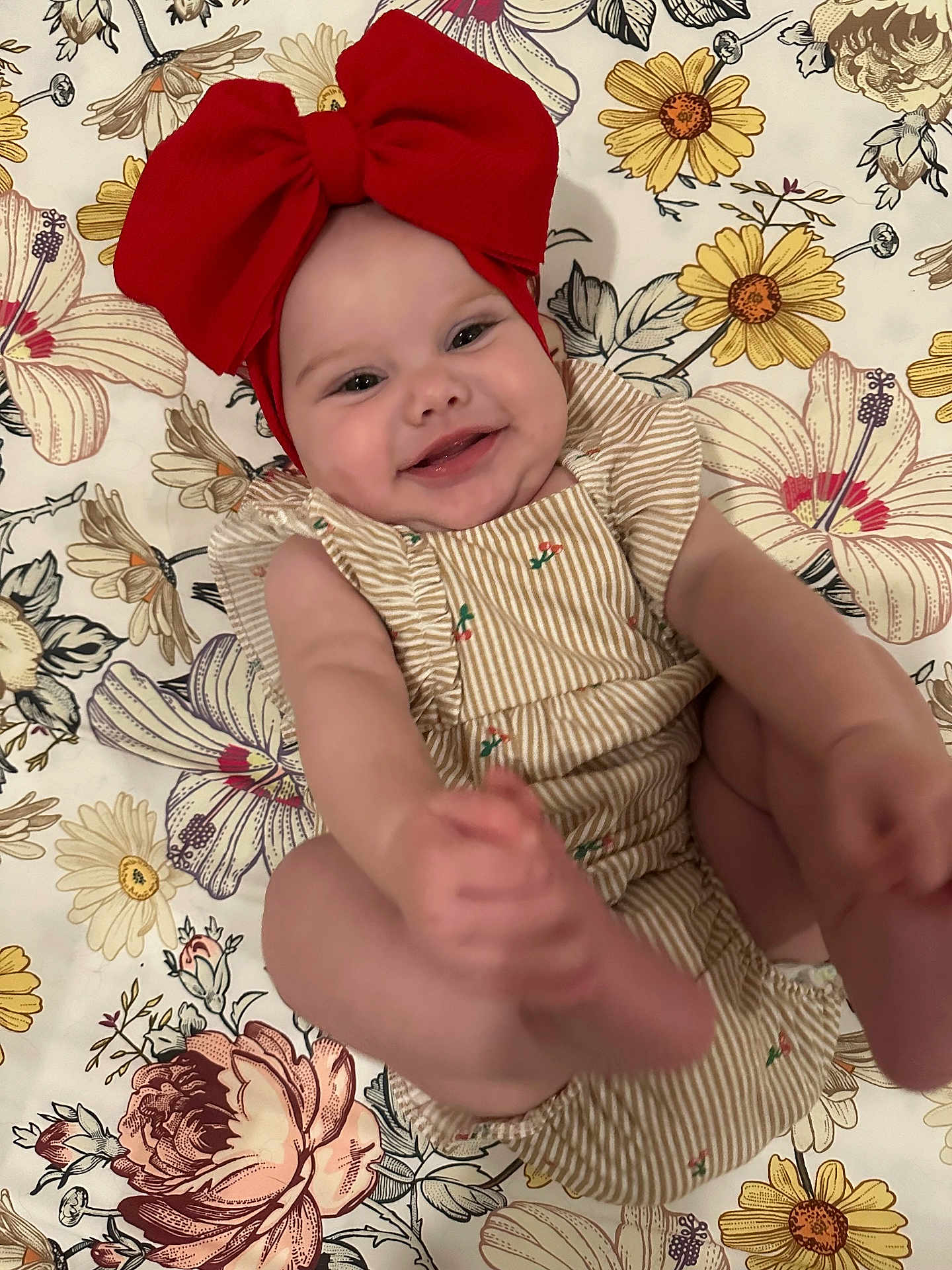 Marymae is registered to the contest to win money with this photo: baby, smiling, red_bow, headband, striped_clothing, floral_pattern, blanket, happy, infant, cute, face, feet, hands, portrait, child, playful, lying_down, skin, cheeks, indoors