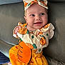 baby, child, smiling, cute, carrot_outfit, headband, orange, yellow, dress, leather_couch, happy, infant, portrait, clothing, sitting, young_child, indoors, face, person, adorable