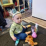 Devlin is registered to the contest to win money with this photo: baby, child, toy, plush_toy, carpet, headband, indoor, floor, clothing, sitting, pacifier, furniture, shelf, chair, person, blue_pants, green_shirt, orange_socks, stuffed_animal, playtime