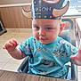 Devlin is registered to the contest to win money with this photo: toddler, child, crown, paper_crown, high_chair, blue_shirt, indoor, table, curious, smiling, person, young_child, light_skin, short_hair, daylight, window, wooden_table, restaurant, seated, cute