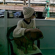 Mickey joined the competition — help win amazing prizes! person, dog, knitted_sweater, knitted_hat, chair, water, dock, rope, petting, sitting, outdoor, brown_dog, green_harness, calm, relaxed, winter_clothing, watercraft, shade, glasses, side_profile