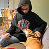Mickey joined the competition — help win amazing prizes! person, dog, hoodie, glasses, couch, indoor, living_room, wooden_chair, window_blinds, casual_clothing, pet, affection, brown_dog, seated, home_interior, furniture, relaxation, companion, looking_down, cozy