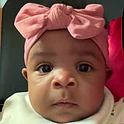 Blessynn is registered to the contest to win money with this photo: baby, infant, newborn, face, eyes, cheeks, headband, bow, pink_bow, bib, close_up, portrait, indoor, pillow, dark_background, expression, curious, adorable, skin, clothing