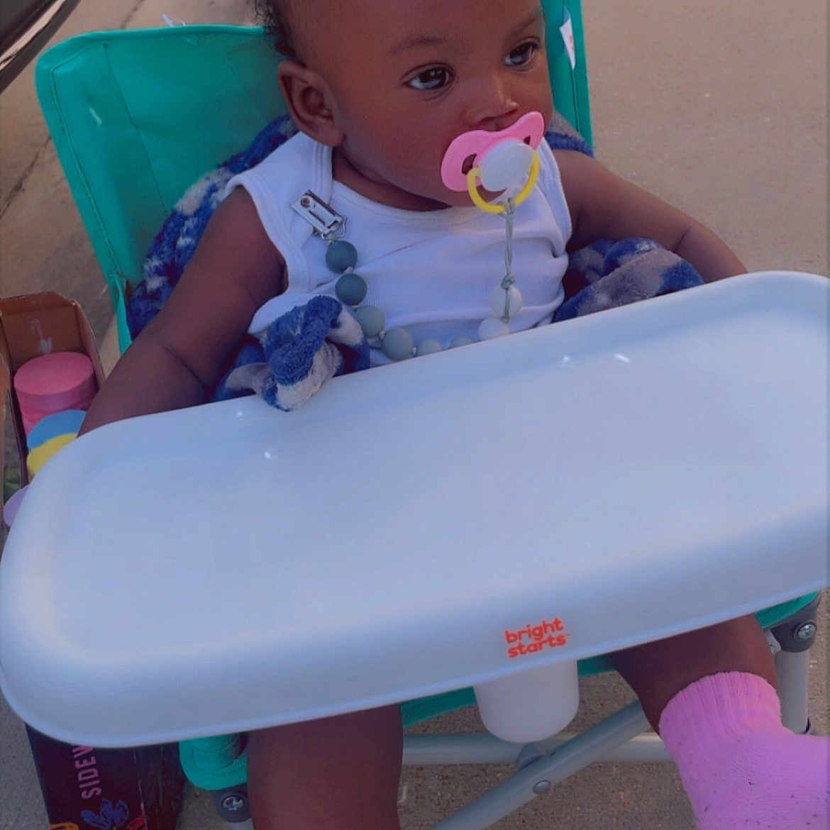 Lazaharia Roddie is registered to the contest to win money with this photo: baby, box, car, casual, child, curly_hair, cute, daylight, high_chair, infant, looking_away, outdoor, pacifier, person, pink_socks, relaxed, seat, sidewalk, toy, white_clothing