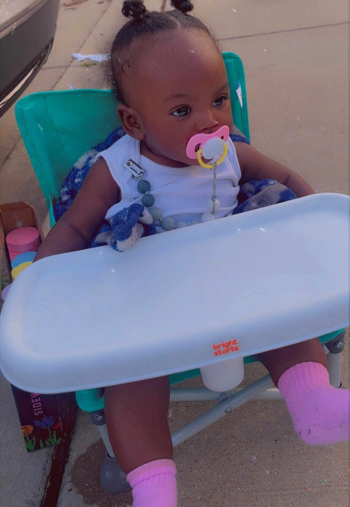 Lazaharia Roddie is registered to the contest to win money with this photo: baby, box, car, casual, child, curly_hair, cute, daylight, high_chair, infant, looking_away, outdoor, pacifier, person, pink_socks, relaxed, seat, sidewalk, toy, white_clothing