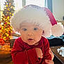 baby, child, santa_hat, christmas, holiday, festive, red_clothing, indoor, portrait, cute, wide_eyes, hand, face, soft_light, decorations, tree, blurred_background, warm, cozy, celebration