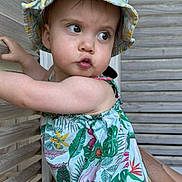 Louise a rejoint le concours — aidez-le/la à gagner de superbes lots ! toddler, child, baby, hat, floral, dress, outdoor, wooden_wall, curious, pouting, person, skin, arm, summer, sunhat, colorful, nature_pattern, portrait, support, casual