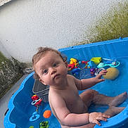 Ilyan a rejoint le concours — aidez-le/la à gagner de superbes lots ! toddler, child, pool, water, toys, blue_pool, bath_toys, curly_hair, wet_skin, outdoor, concrete_floor, moss, plastic, sitting, playtime, summer, bath, baby, cute, curious