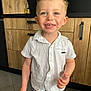 toddler, child, smiling, blue_eyes, blonde_hair, white_shirt, floral_shorts, pacifier, kitchen, wooden_cabinets, modern_interior, happy, standing, person, indoor, cute, portrait, casual_clothing, young_child, light