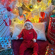 Michael is registered to the contest to win money with this photo: baby, child, christmas_tree, christmas_decorations, presents, gift_bags, red_clothing, santa_hat, sneakers, holiday, festive, smiling, indoors, cushion, white_tree, ornaments, butterfly_decoration, lighting, happy, celebration