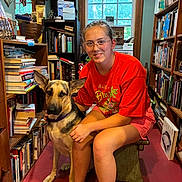 Mindy is registered to the contest to win money with this photo: person, dog, german_shepherd, bookshelf, books, indoor, bench, window, glasses, red_shirt, shorts, sitting, pet, animal, floor, wooden_furniture, smile, casual_clothing, home, reading