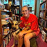 person, dog, german_shepherd, bookshelf, books, indoor, bench, window, glasses, red_shirt, shorts, sitting, pet, animal, floor, wooden_furniture, smile, casual_clothing, home, reading