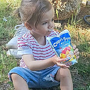 Illanna a rejoint le concours — aidez-le/la à gagner de superbes lots ! adult, capri_sun, casual_clothing, child, daylight, drink_pouch, drinking, grass, greenery, legs, nature, outdoor, person, pink_shoes, shorts, sitting, sneakers, stone_seat, striped_shirt, sunlight