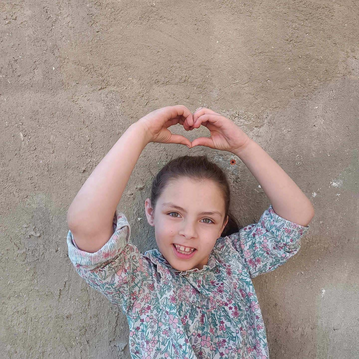 Lyhana a rejoint le concours — aidez-le/la à gagner de superbes lots ! arm, casual_clothing, child, cute, face, floral_shirt, girl, hands, happy, heart_shape, human, natural_light, outdoor, portrait, posing, skin, smile, texture, wall, young