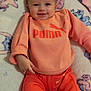 baby, smiling, infant, orange_clothing, puma_logo, blanket, soft_texture, cute, happy, child, portrait, indoor, sitting, cozy, warm_clothing, face, person, fashion, casual, comfort