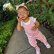 Aliyah is registered to the contest to win money with this photo: toddler, child, glasses, headband, pink_clothing, overalls, sneakers, sidewalk, outdoor, greenery, plants, fence, person, portrait, young_child, casual_wear, nature, daylight, cute, fashion