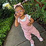 toddler, child, glasses, headband, pink_clothing, overalls, sneakers, sidewalk, outdoor, greenery, plants, fence, person, portrait, young_child, casual_wear, nature, daylight, cute, fashion