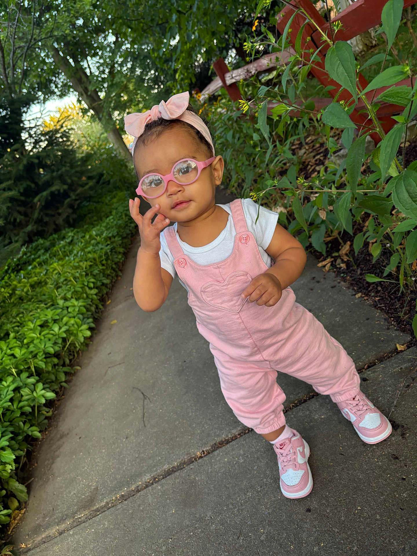 Aliyah is registered to the contest to win money with this photo: toddler, child, glasses, headband, pink_clothing, overalls, sneakers, sidewalk, outdoor, greenery, plants, fence, person, portrait, young_child, casual_wear, nature, daylight, cute, fashion
