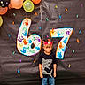 child, boy, birthday, crown, balloons, handprints, numbers, black_background, colorful, celebration, party, smiling, shirt, shark, jeans, standing, decorations, festive, happy, portrait
